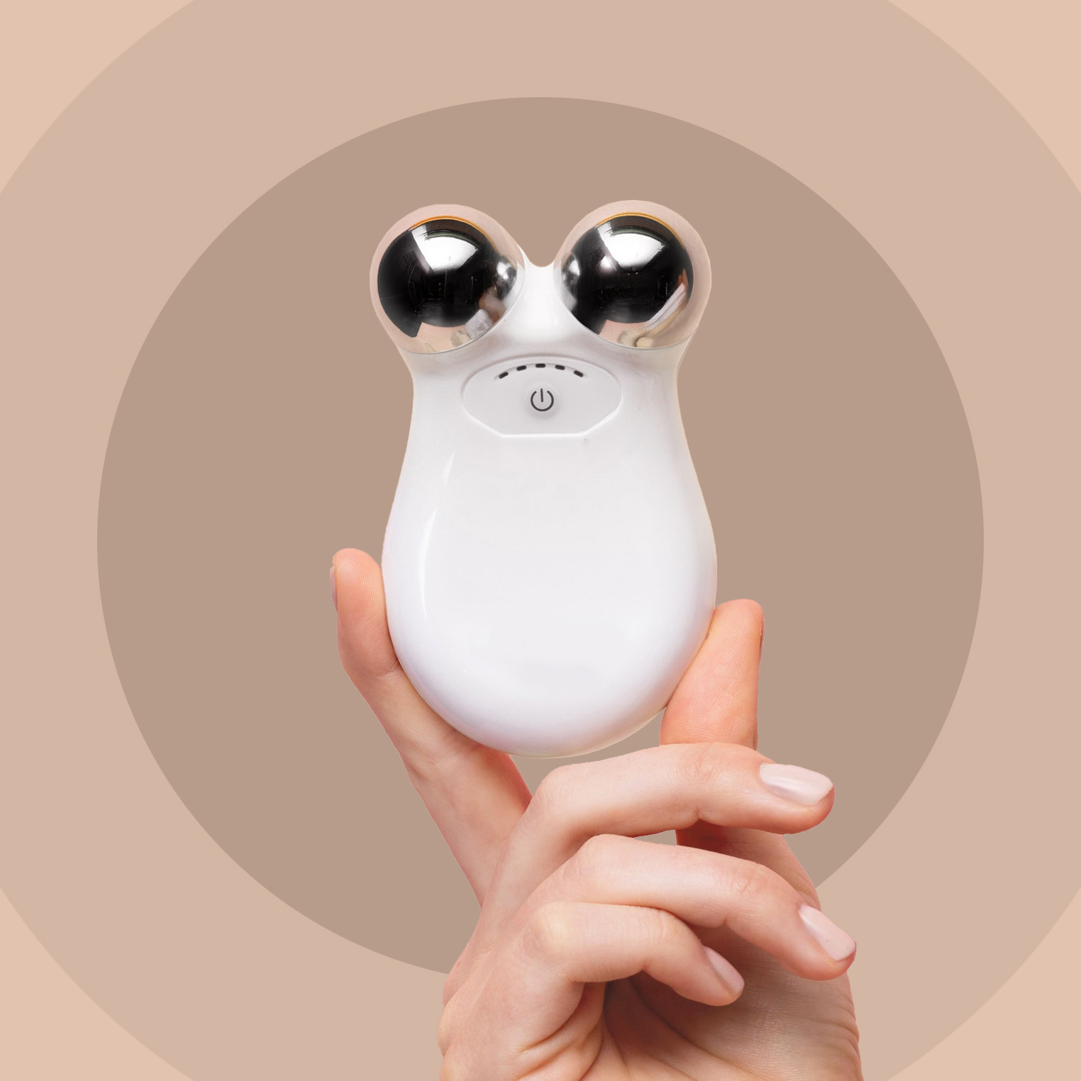 A hand holding a white handheld facial toning device with two silver spheres against a circular brown background.