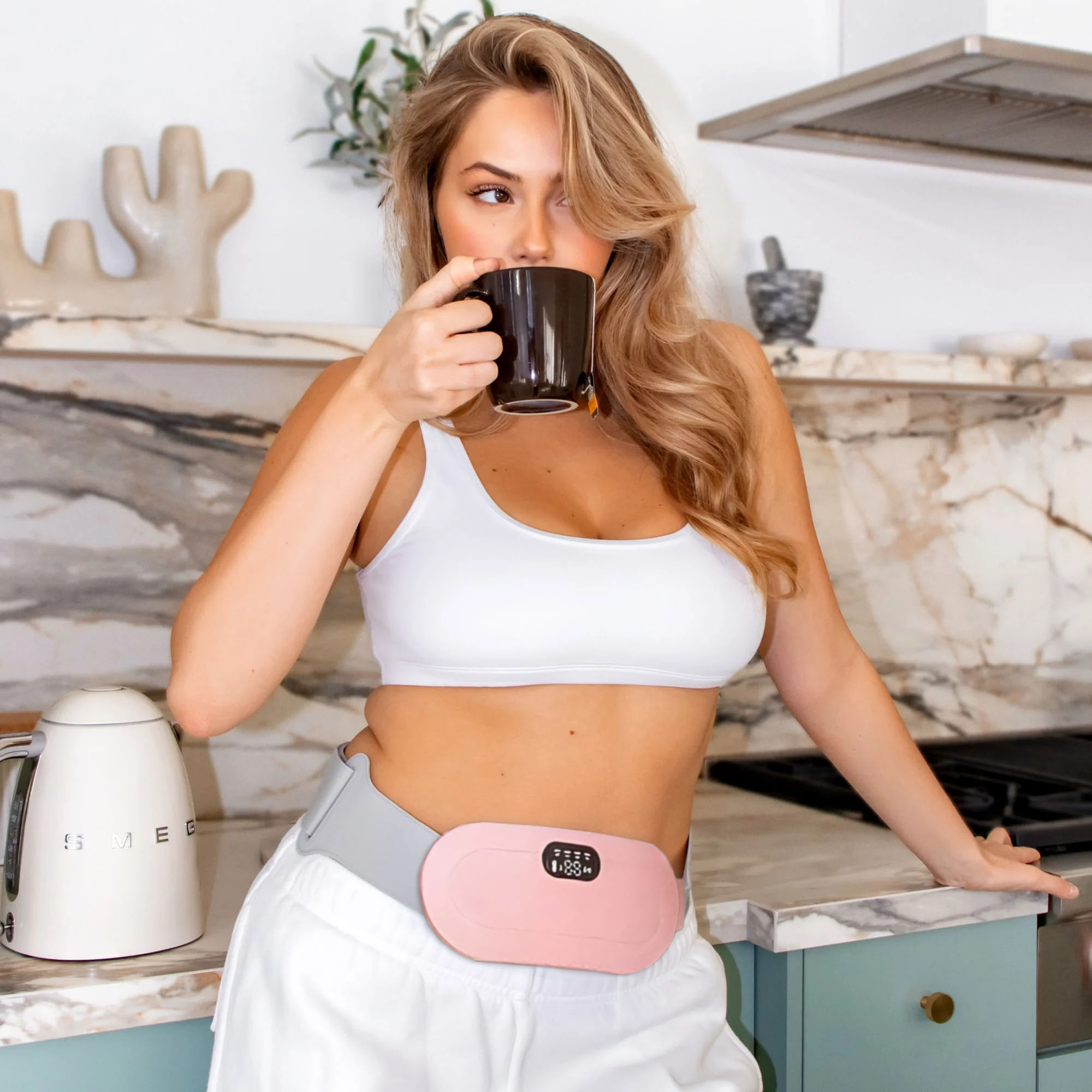 Person in kitchen wearing a white top, holding a mug, with a pink device on waist.