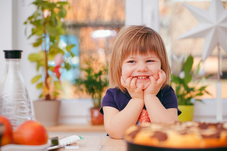 Smiling child seated at a table with plants and a dessert nearby.