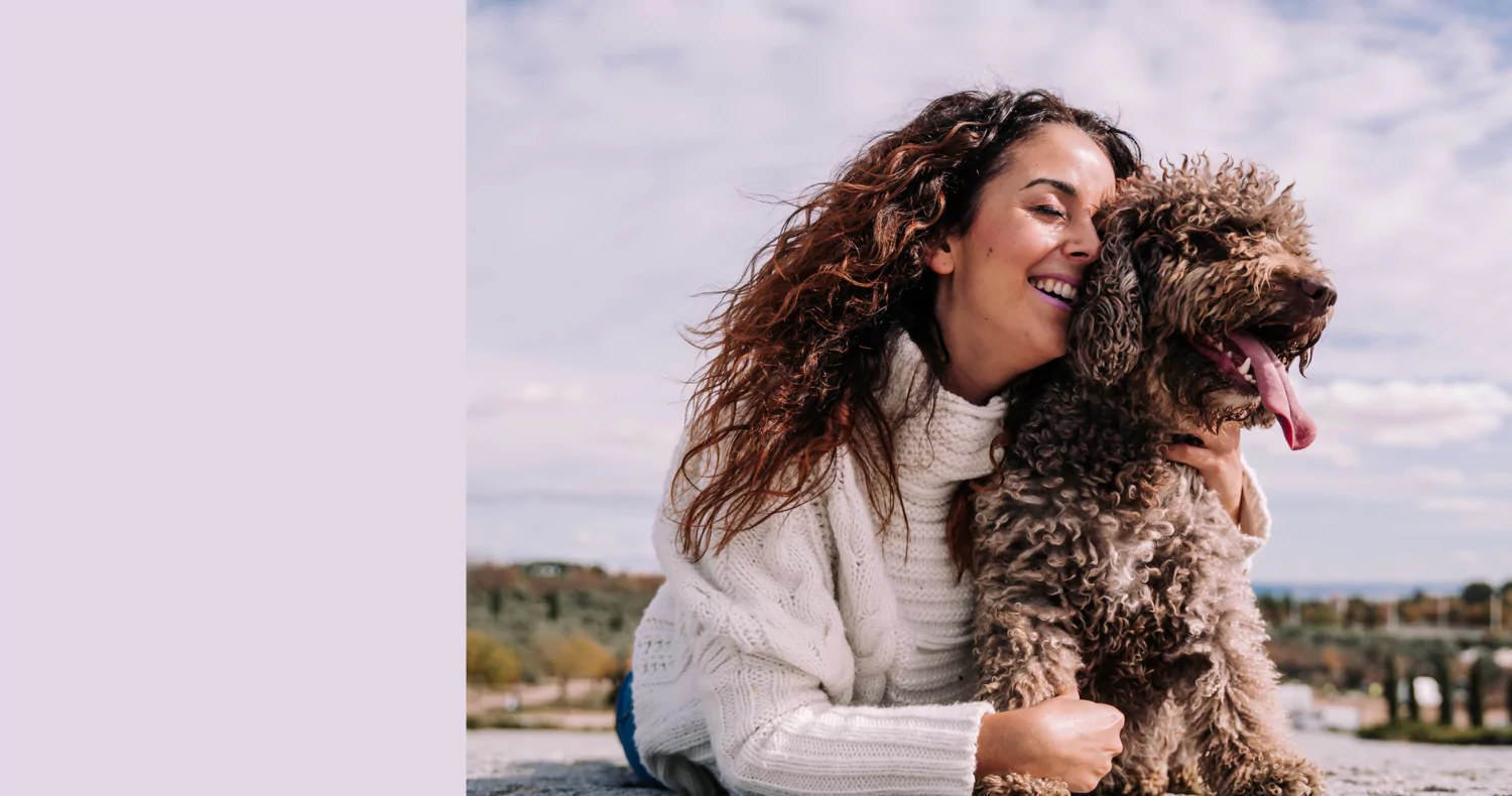 Woman smiling and hugging a curly-haired dog outdoors.