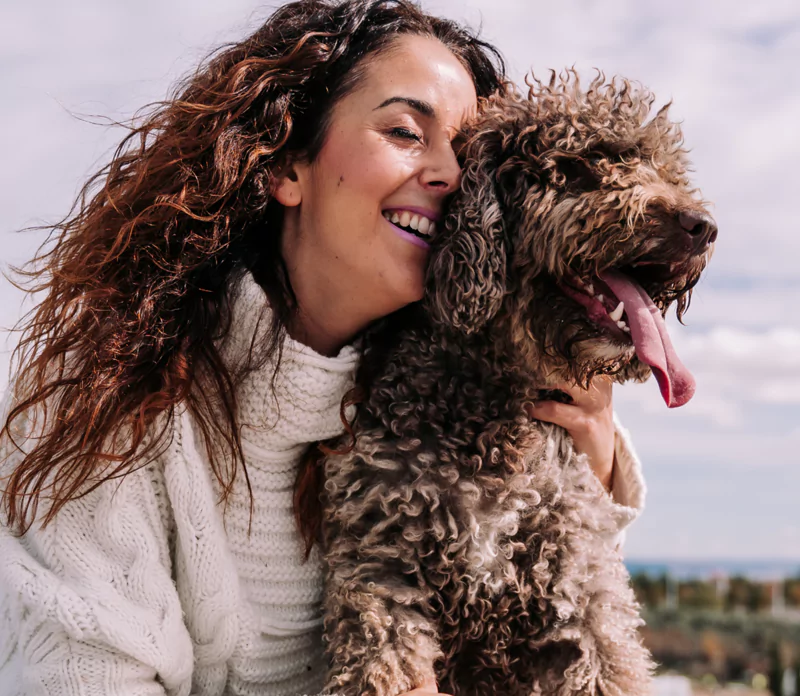 Woman smiling and hugging a curly-haired dog outdoors.