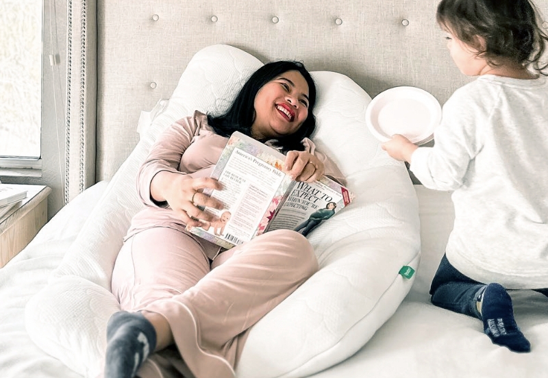 Woman relaxing with The Newton Pillow in bed with a book, smiling at child holding a plate.