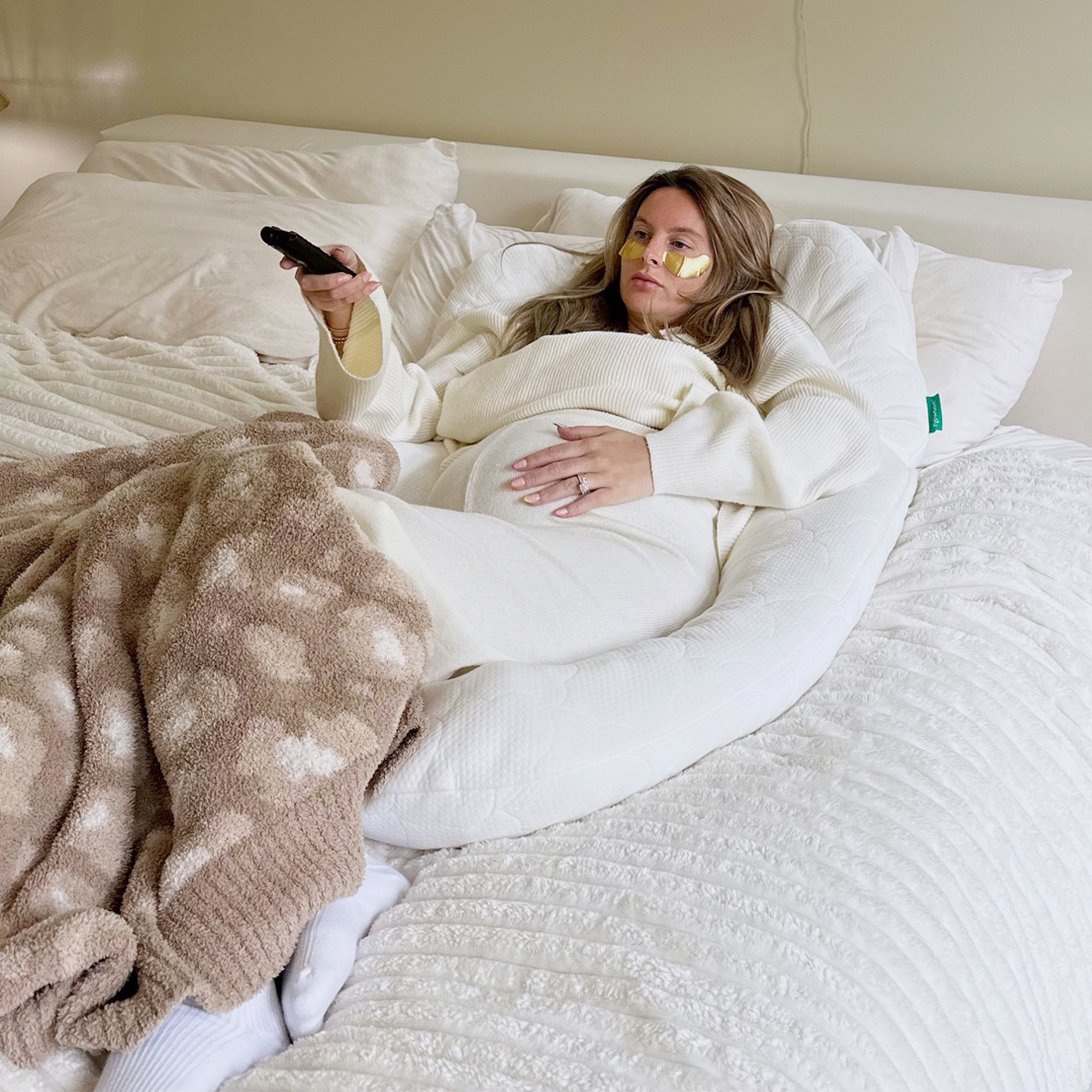 Pregnant woman lounging in bed with The Newton Pillow with TV remote in hand