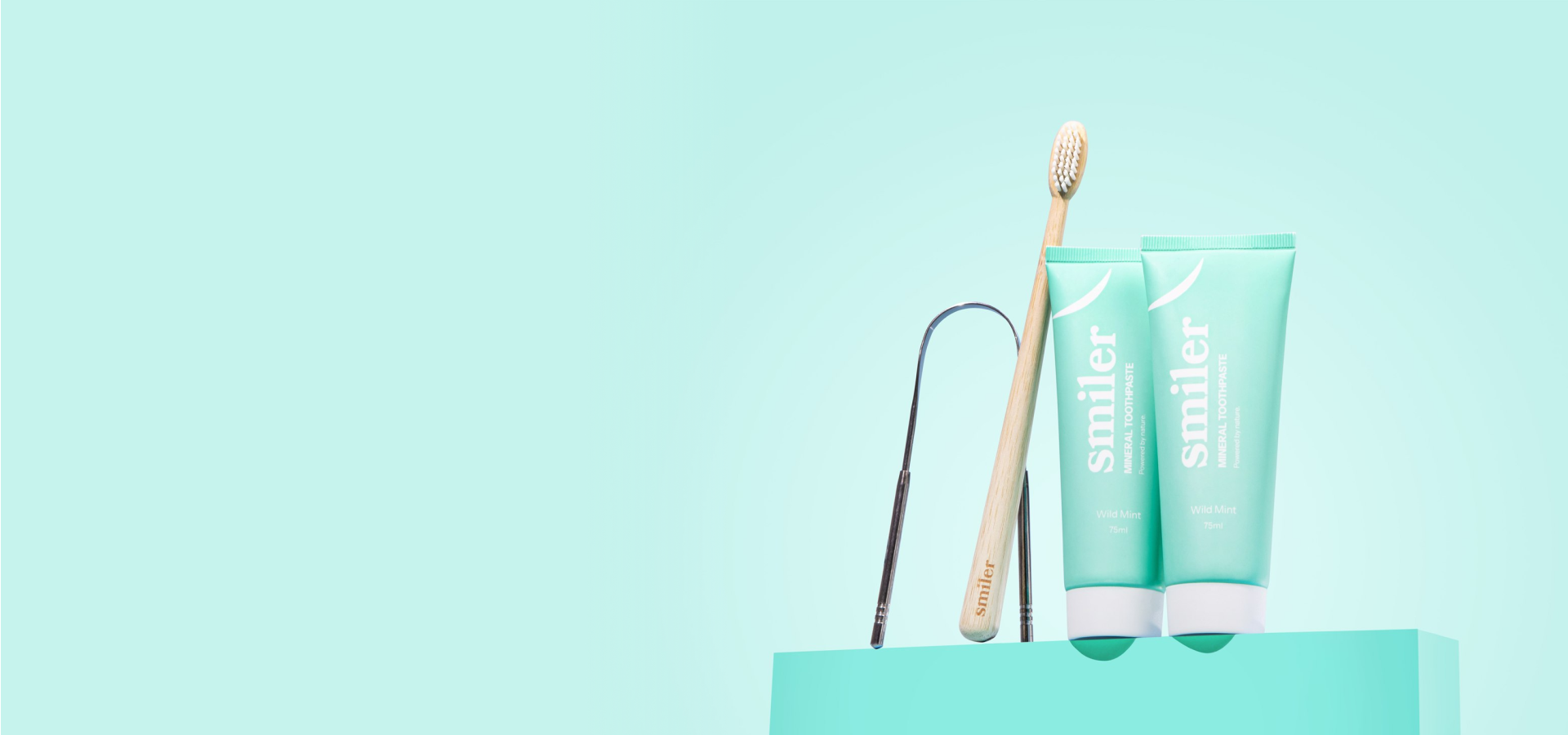 Two tubes of Smiler mineral toothpaste, a wooden toothbrush, and a tongue scraper against a light blue background.