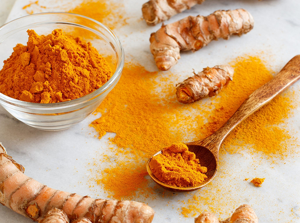 Bowl of turmeric powder with fresh roots and wooden spoon on a surface.