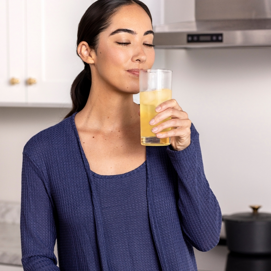 Person in blue attire drinking a glass of iced yellow beverage in a kitchen.