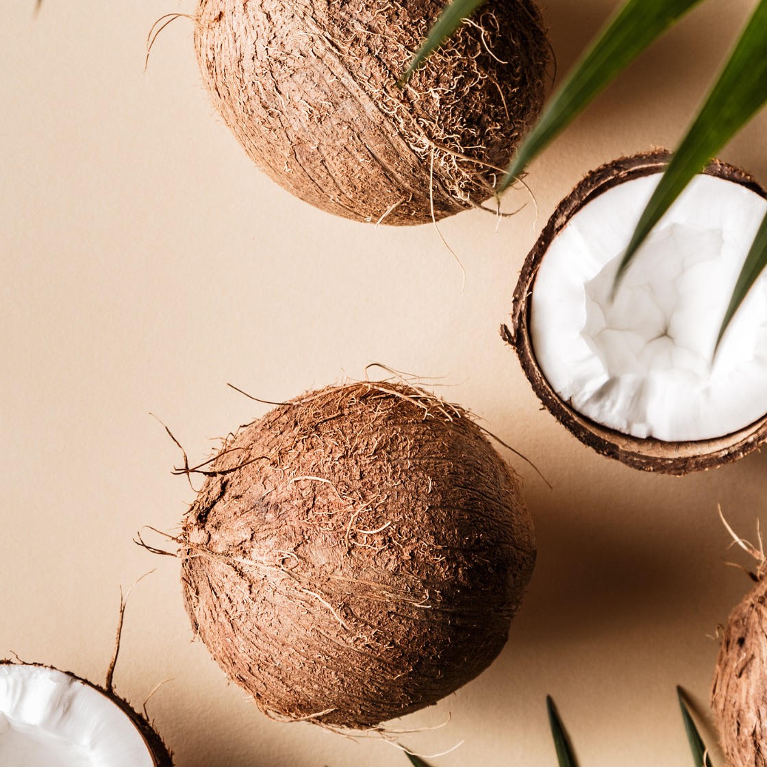 Whole and halved coconuts with green leaves on a beige background.