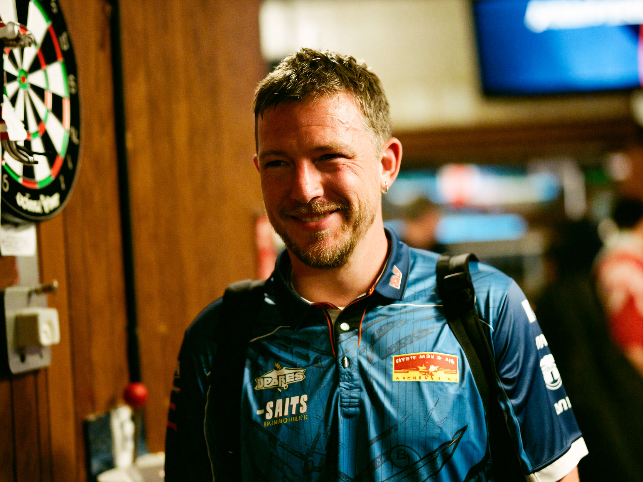 A person in a blue shirt standing indoors near a dartboard.