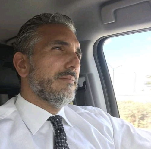 Man with gray hair and beard driving a car, wearing a white shirt and tie.
