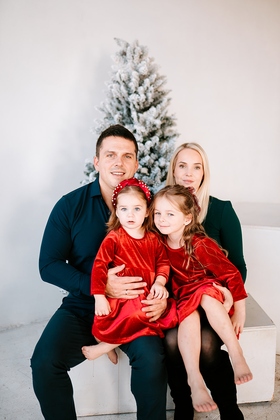 A family of four poses for a Christmas photo, with two young girls in red dresses sitting on their parents' laps.