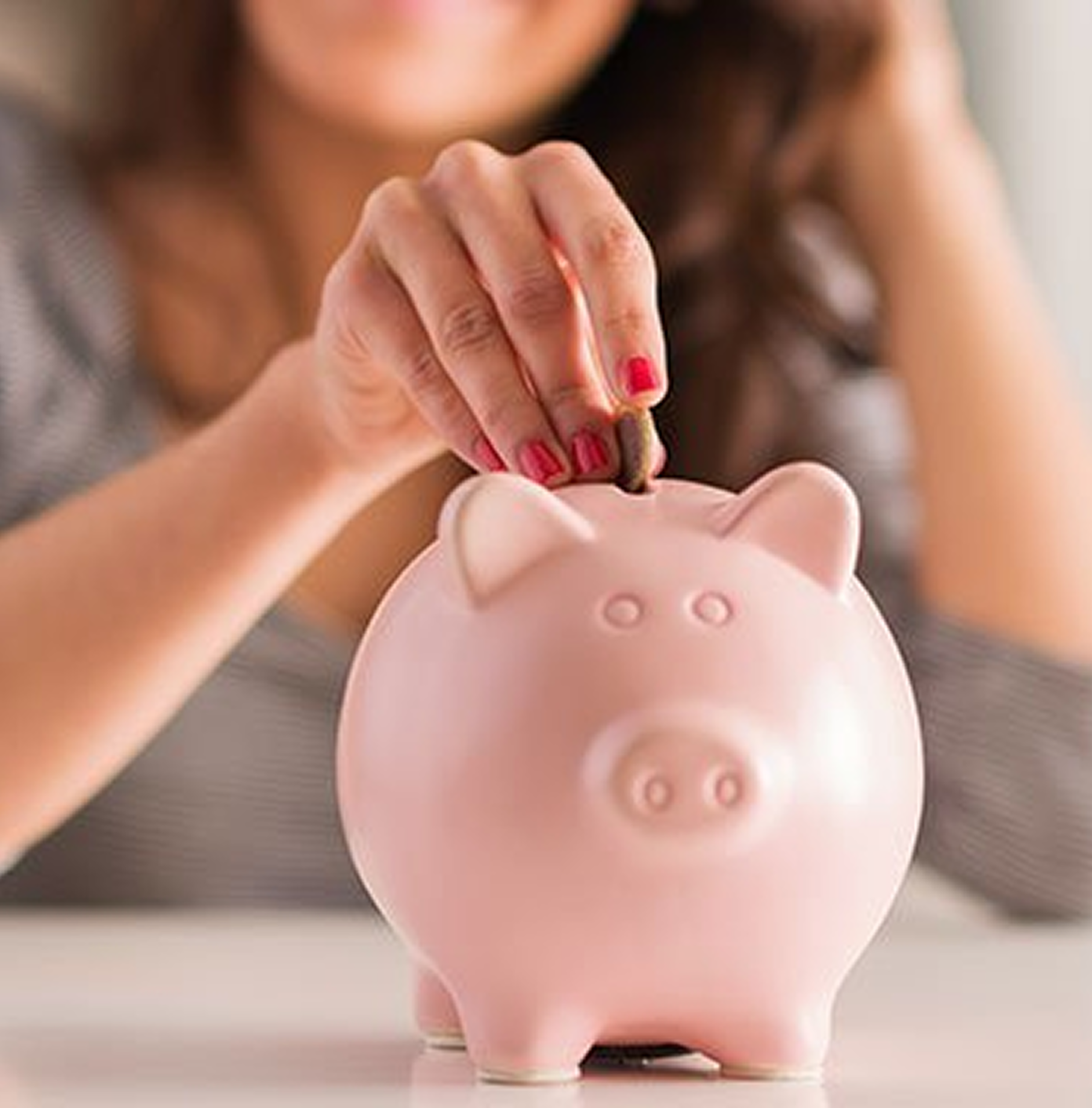 A woman with red nail polish puts a coin into a pink piggy bank on a white table.