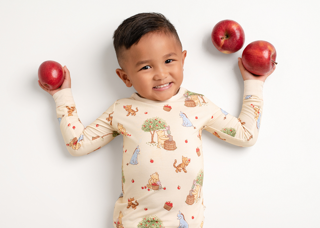 A smiling boy in Winnie the Pooh pajamas lies on a white background holding three red apples.