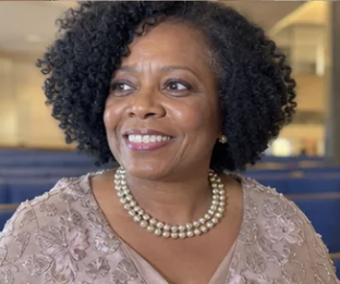 A smiling, middle-aged Black woman with curly hair and a pearl necklace, looking off to the side.