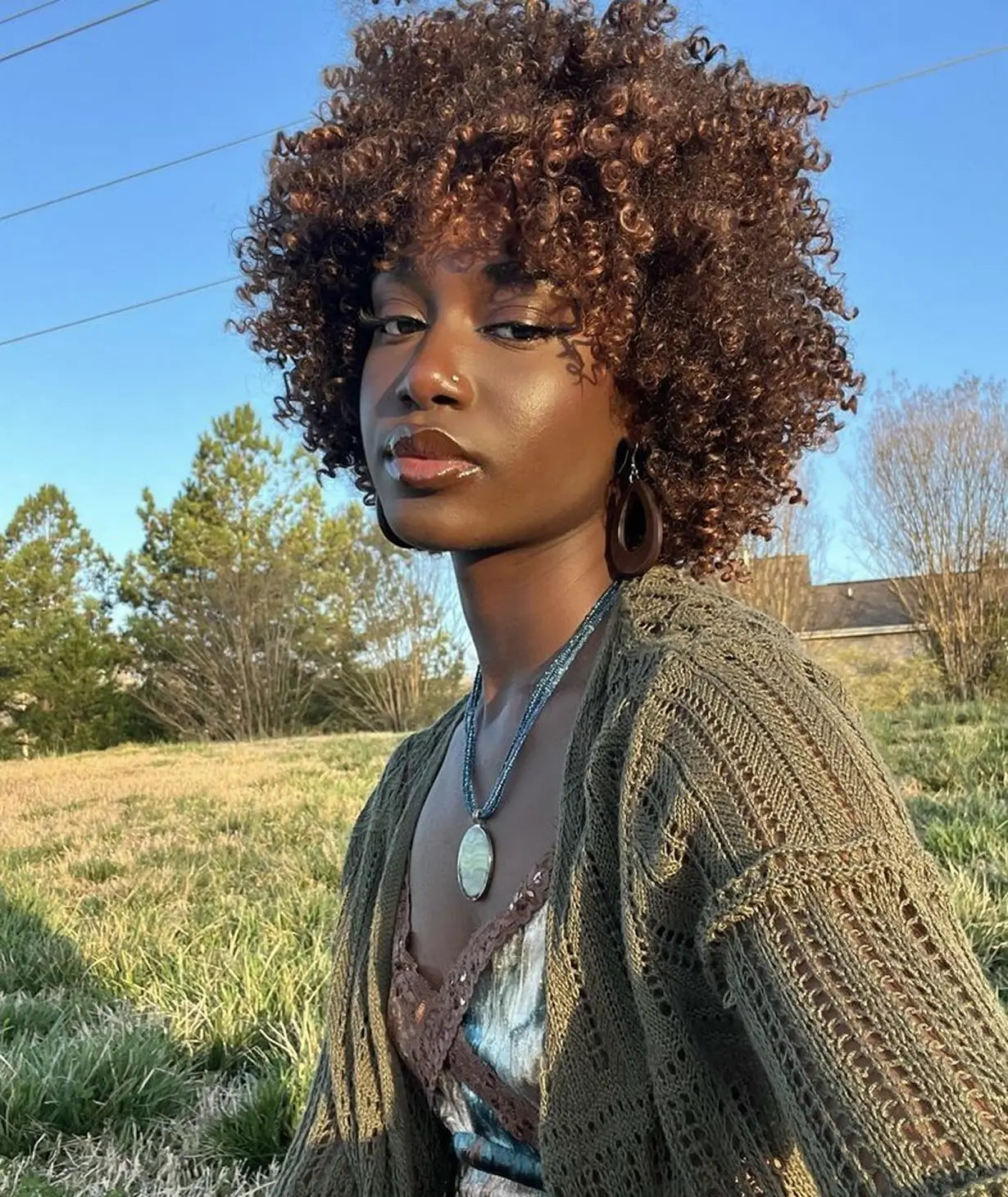 A young woman with curly brown hair poses outside in a grassy field on a sunny day.