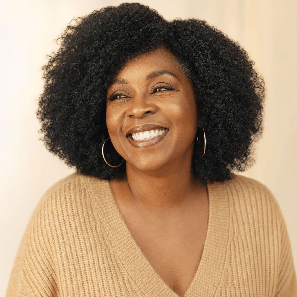 A headshot of a smiling Black woman with curly hair, wearing a tan sweater and gold hoop earrings.