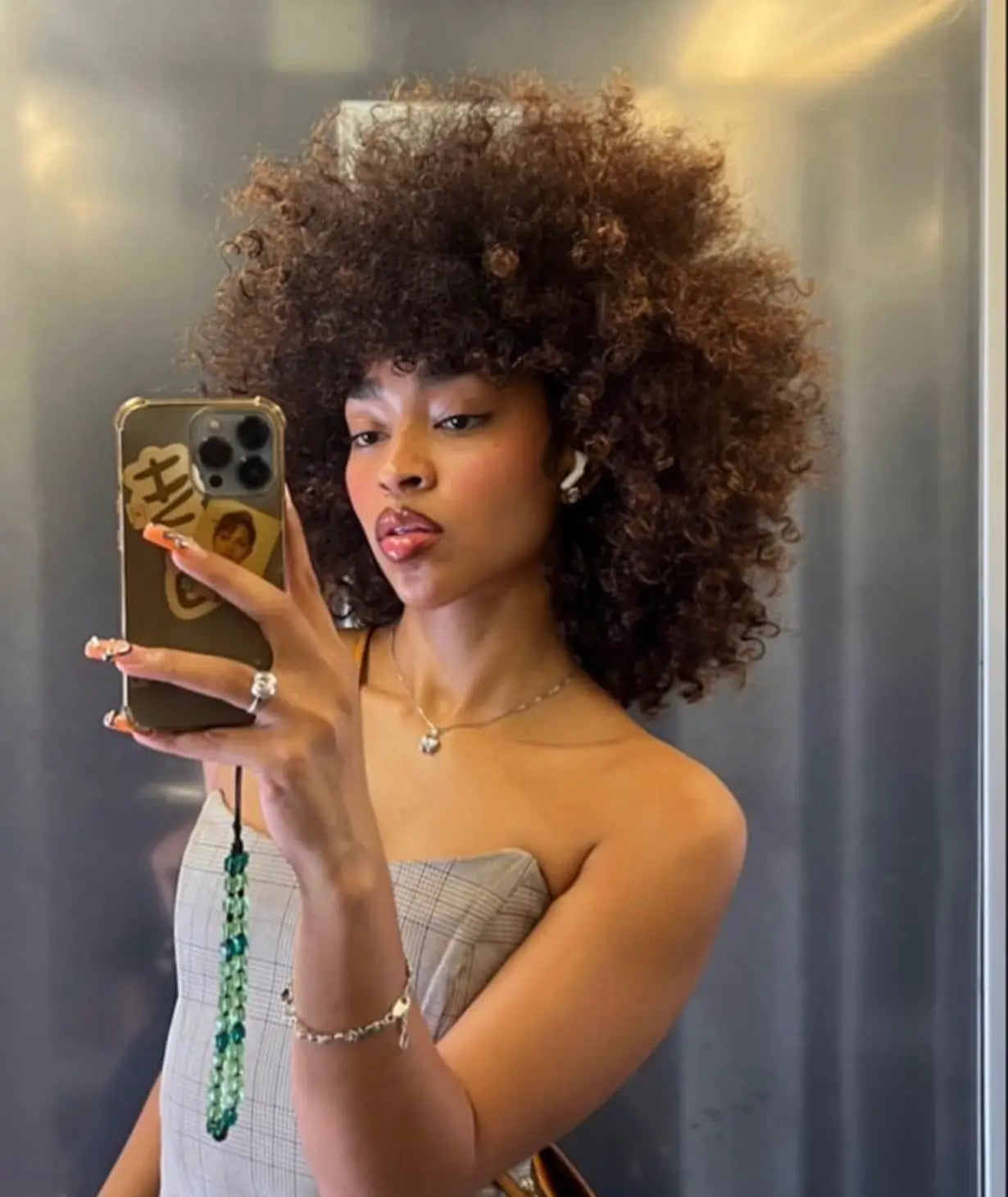 A woman with a large afro hairstyle takes a selfie in a mirror with her smartphone.