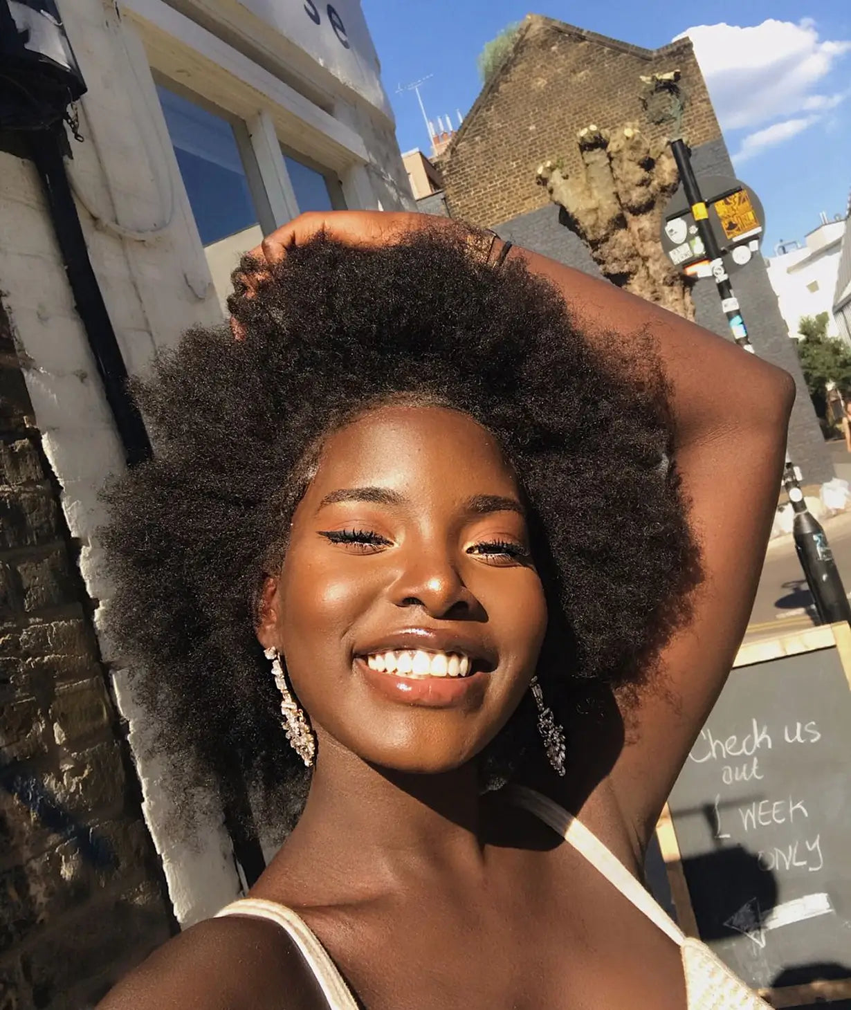A young Black woman with a large afro smiling brightly while standing on a sunny street.