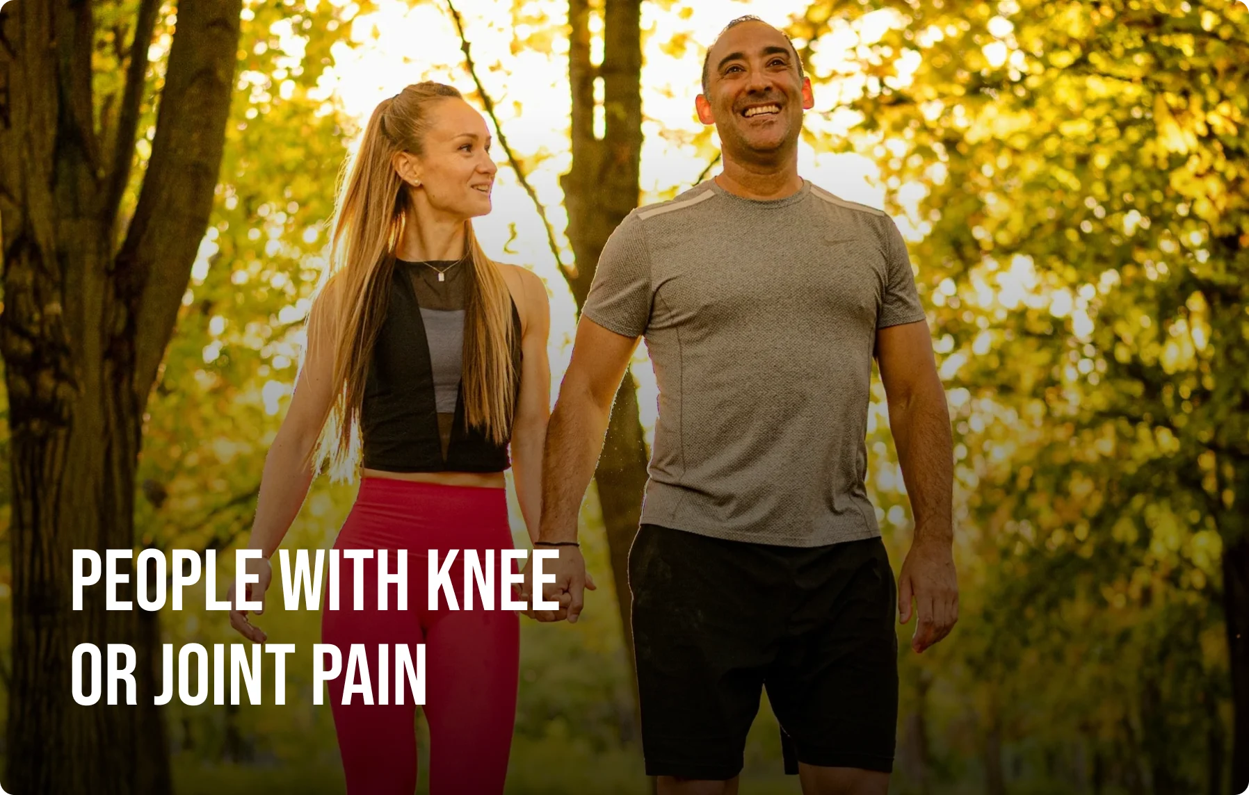 A smiling man and woman in workout clothes hold hands while walking through a sunlit park.