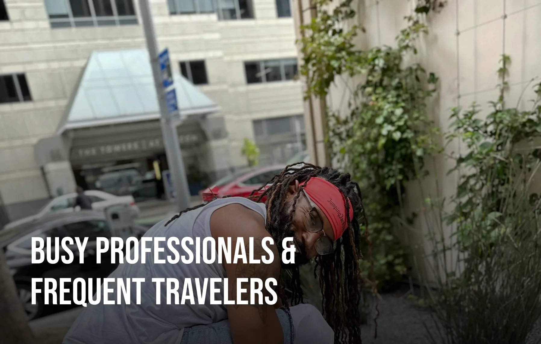 A man with dreadlocks and a red headband looks down while on a city street next to some plants.
