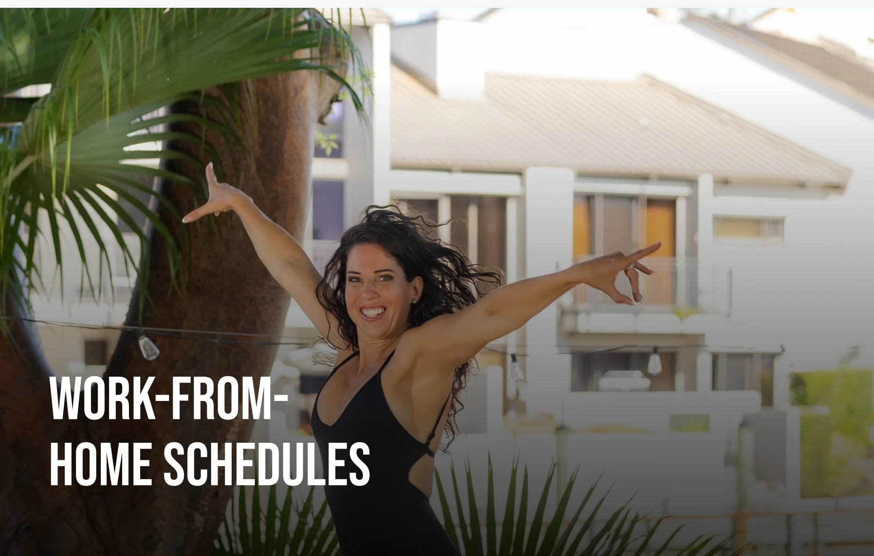 A smiling woman in a black top poses with her arms outstretched outdoors, with palm leaves framing the shot.