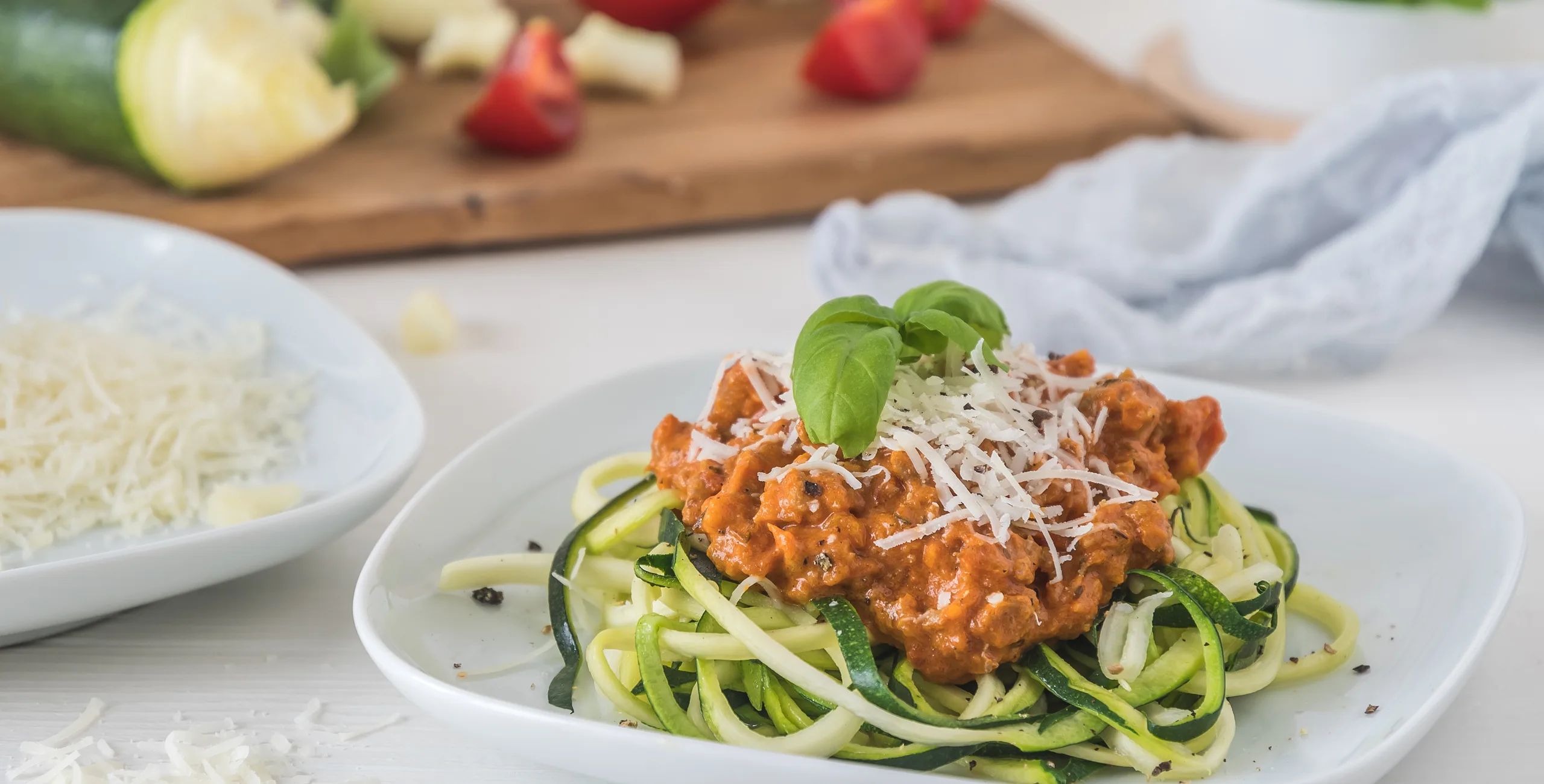 Zucchini noodles with tomato sauce, topped with cheese and basil, on a white plate.