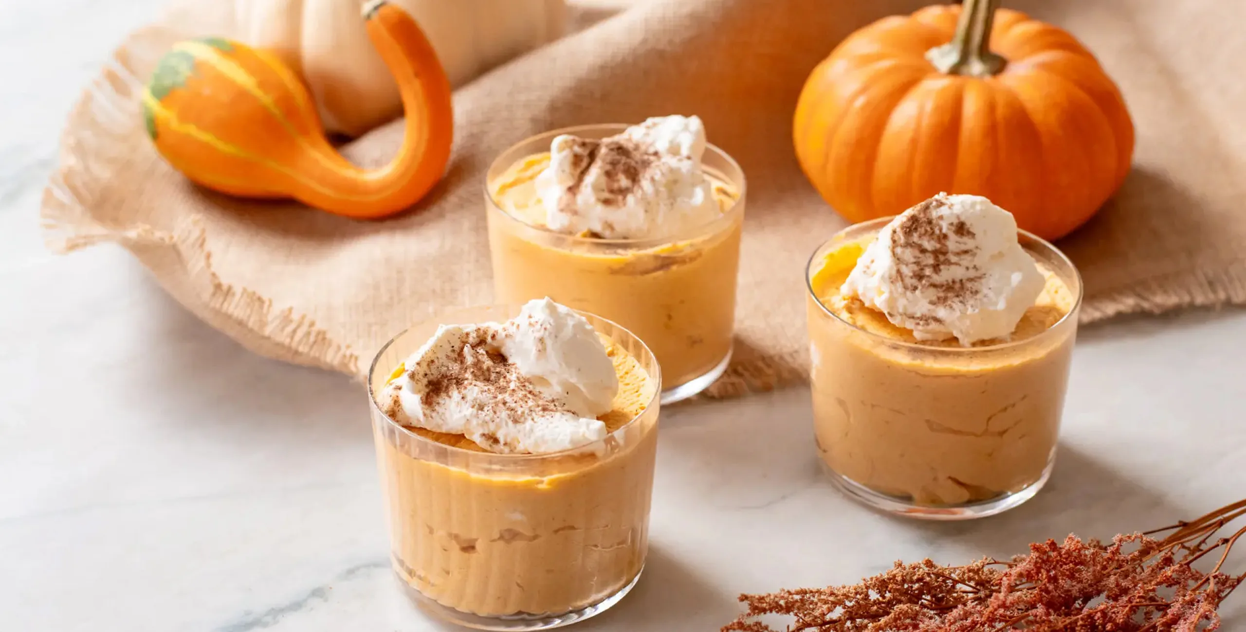 Three glasses of pumpkin mousse with whipped cream and cinnamon, surrounded by pumpkins and gourds.