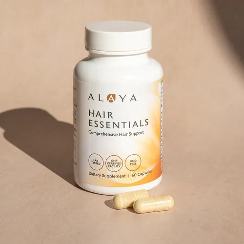A white bottle of Alaya Hair Essentials dietary supplement with two capsules in front of it on a surface.