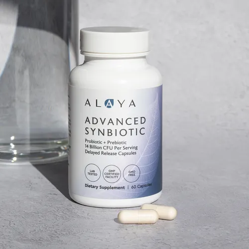 A white bottle of Alaya Advanced Synbiotic supplement with two capsules on a table next to a glass.