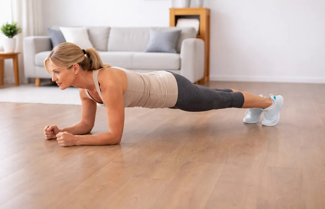 A woman in workout clothes holds a forearm plank position on a wooden floor in a living room.