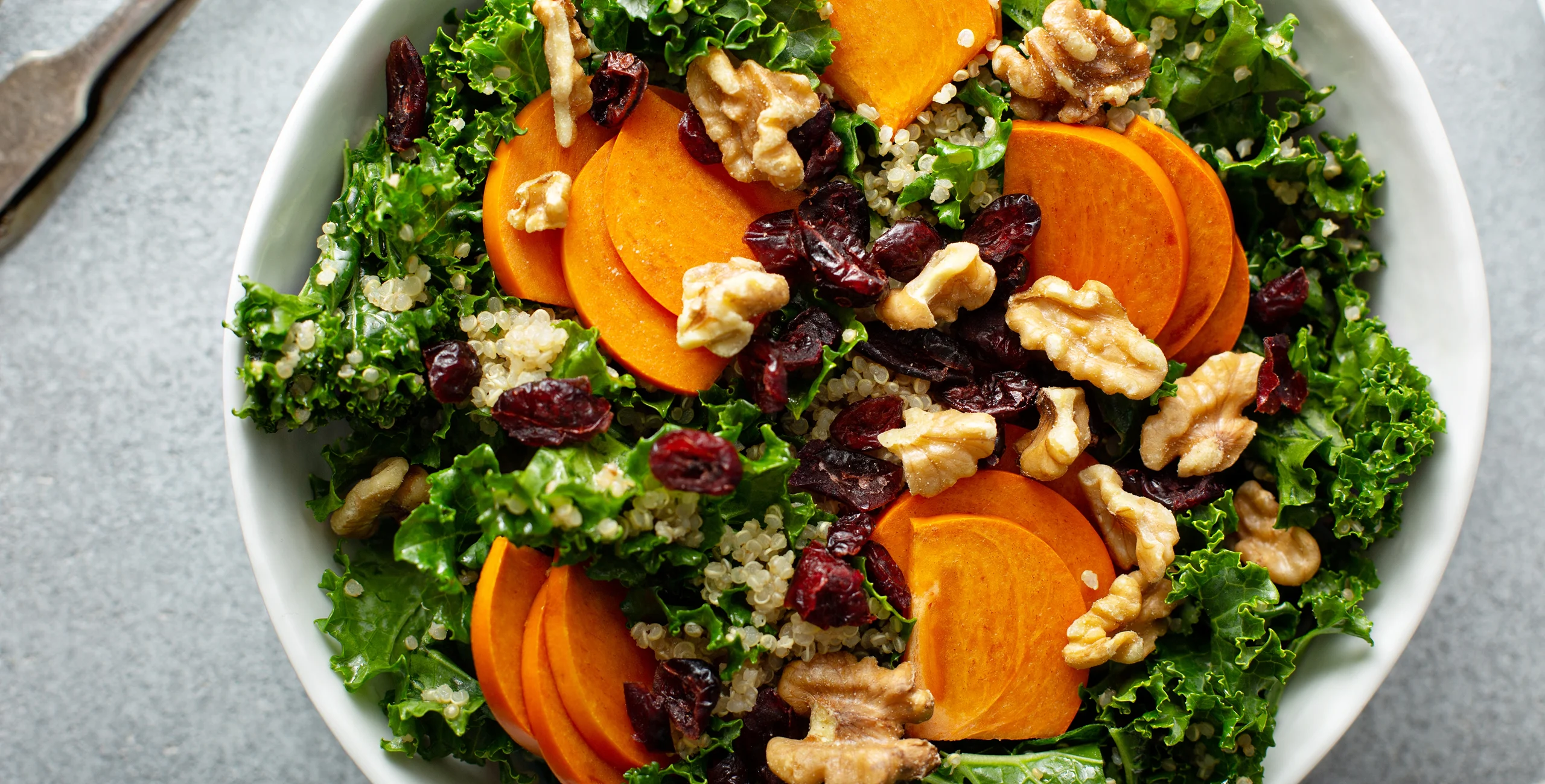 Salad with kale, quinoa, persimmon slices, walnuts, and dried cranberries in a white bowl.