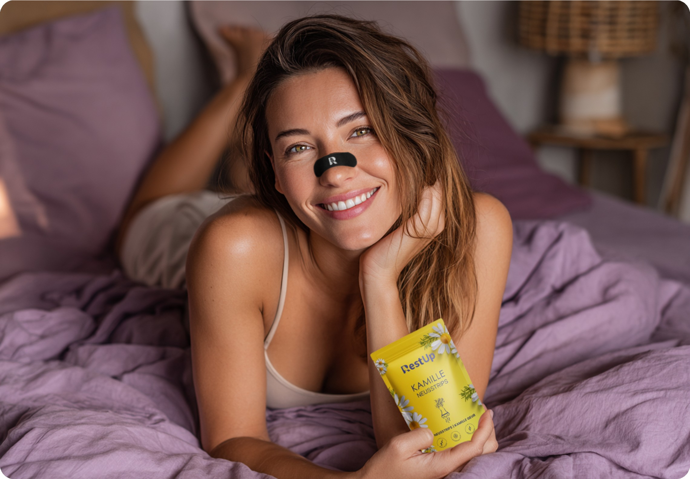 Smiling person with a nose strip, holding a yellow packet, lying on a bed with purple sheets.