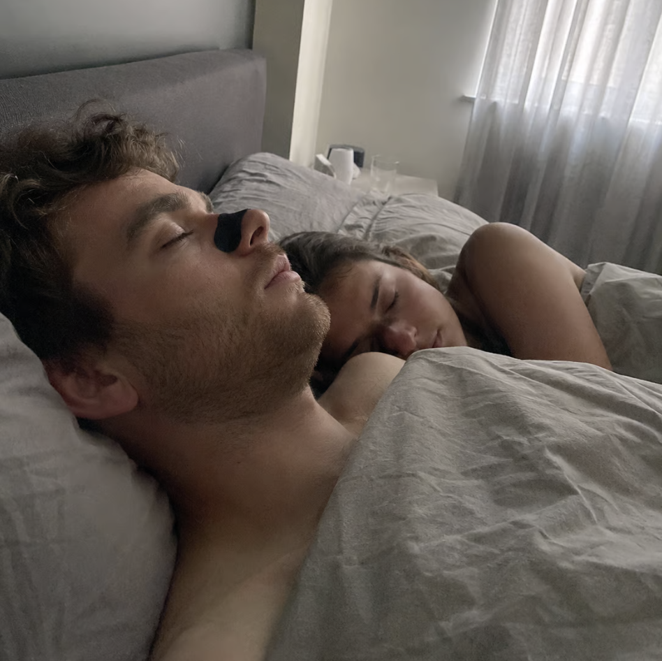 A man wearing a nasal strip and a woman sleeping soundly next to each other in a bed.