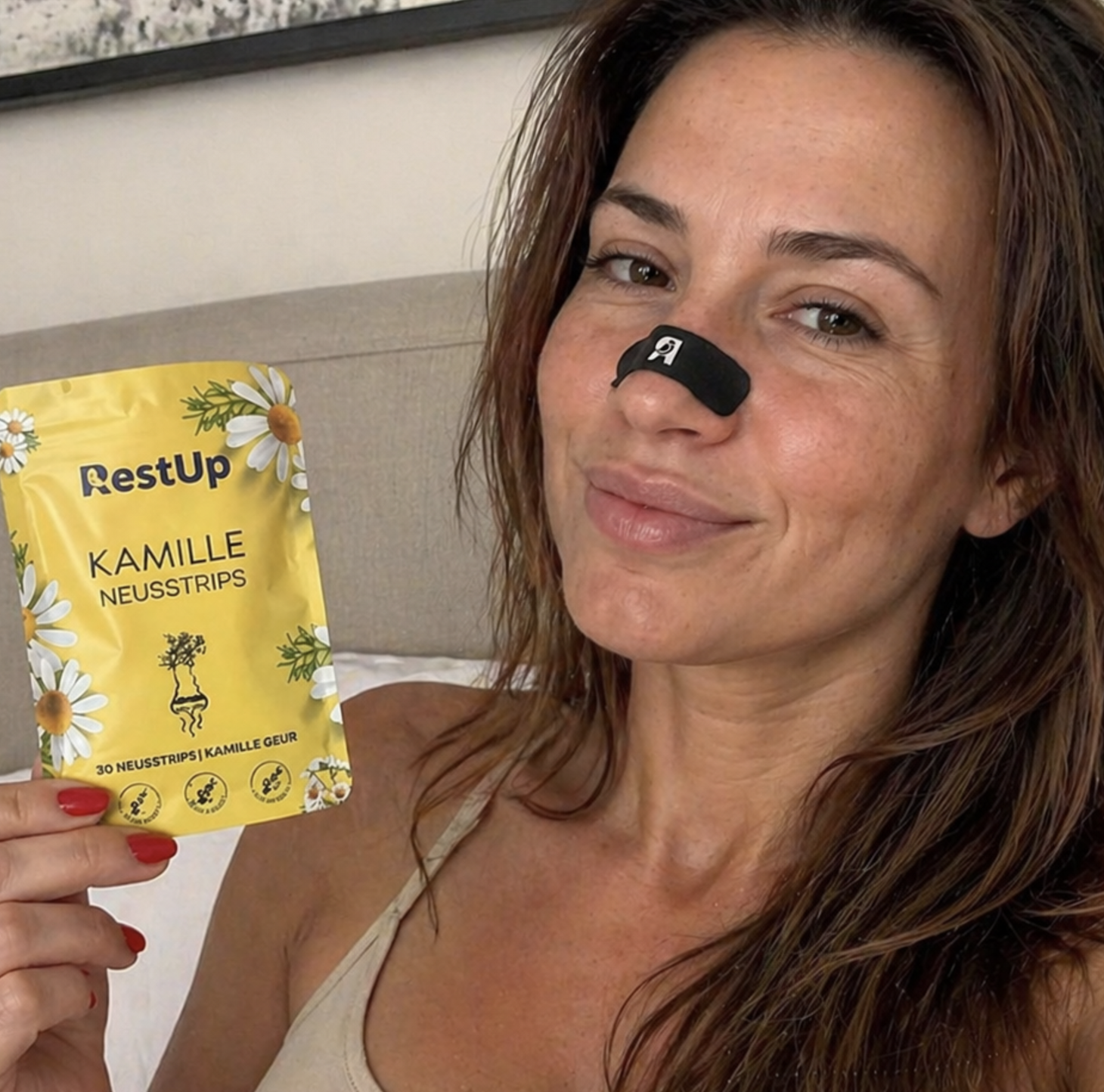 A woman with a black strip on her nose holds a yellow pouch of RestUp Kamille Neusstrips.