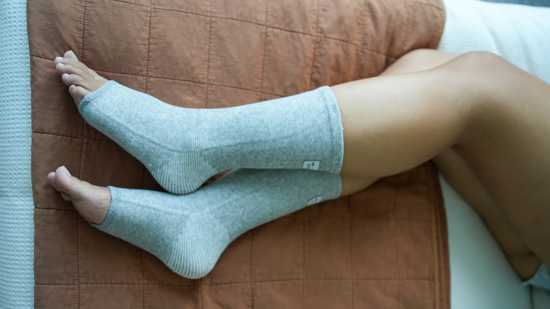 A person's lower legs and feet wearing grey, open-toed socks, resting on a brown quilted surface.