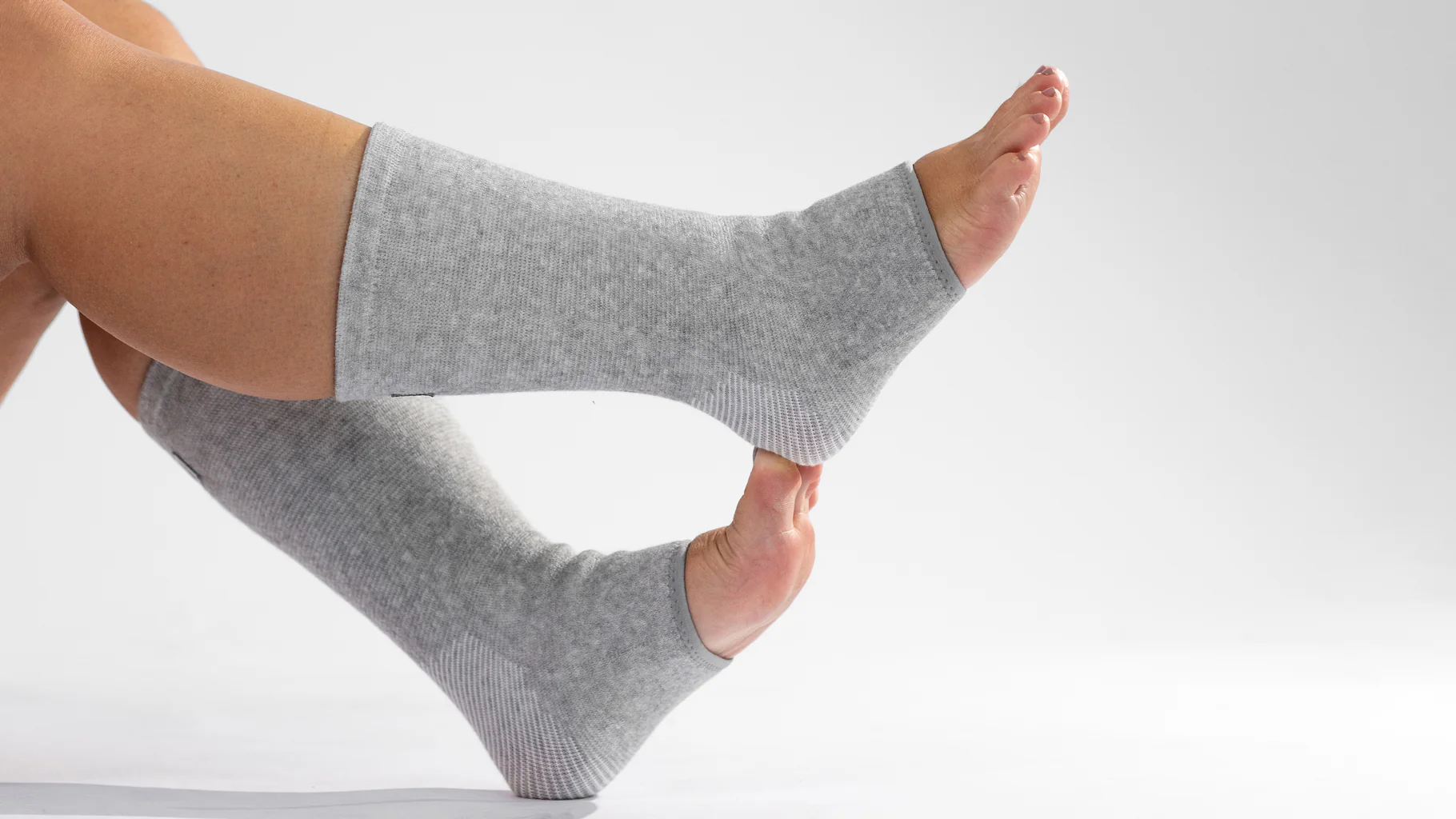 A person's legs and pointed feet wearing grey open-toed and open-heeled socks against a white background.