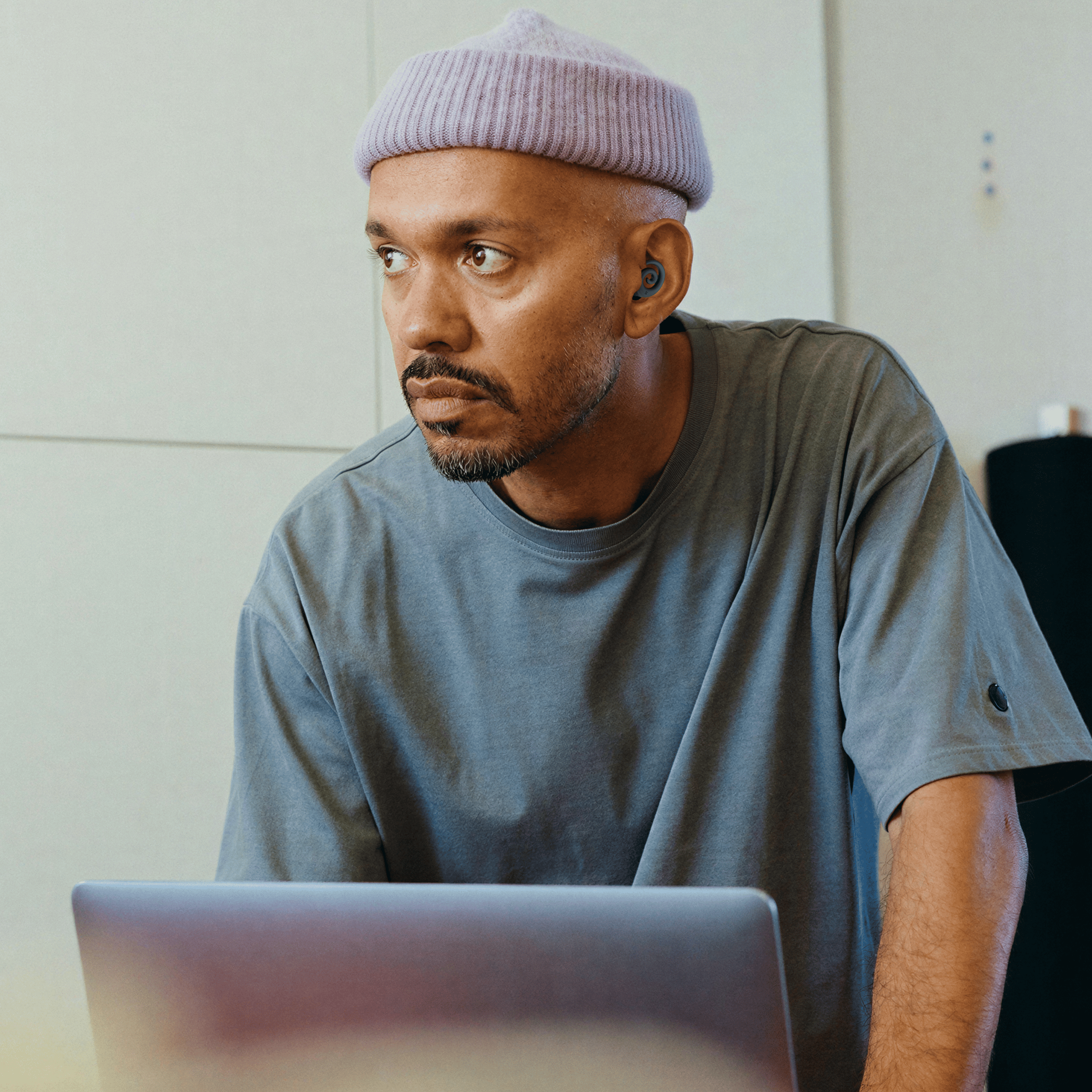 A man in a lavender beanie and an earbud stands behind a laptop, looking off to the side.