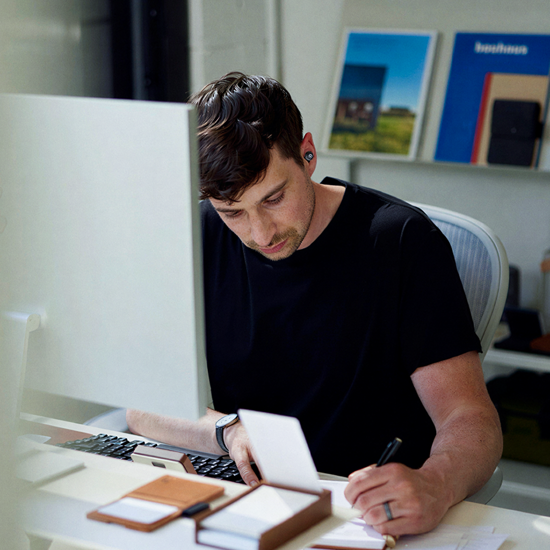 A man wearing an earbud sits at a desk, looking down while writing in a notebook.