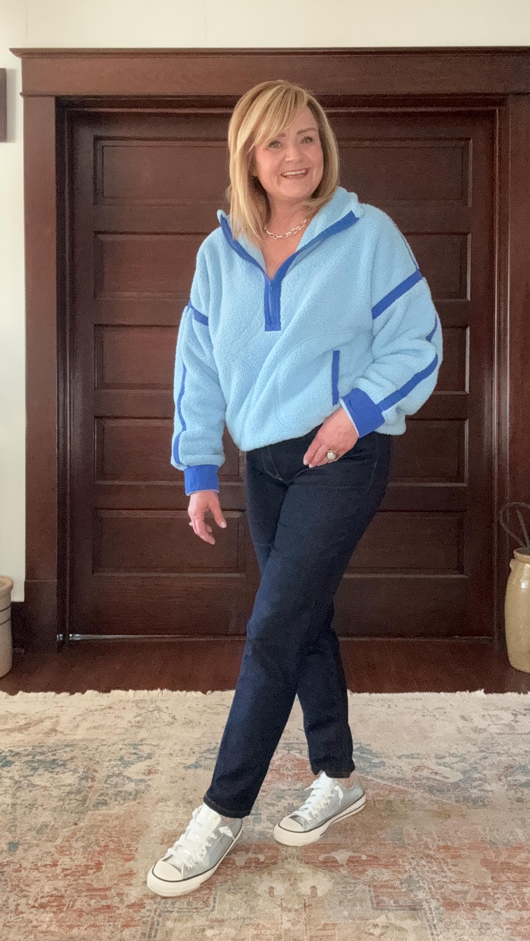 Person in a blue sweater and jeans standing indoors, smiling.