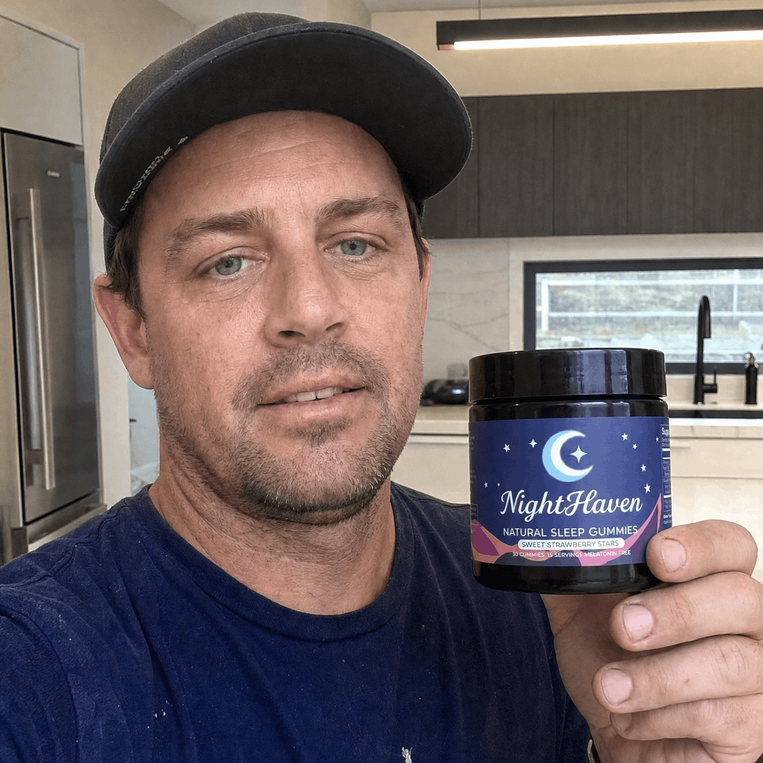 A man in a baseball cap holds up a jar of NightHaven Natural Sleep Gummies in a kitchen.