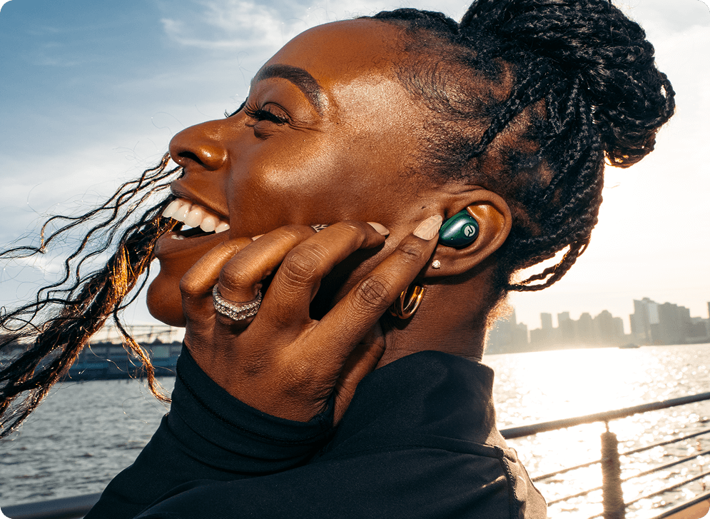 Smiling person with earbuds, standing by a waterfront at sunset.