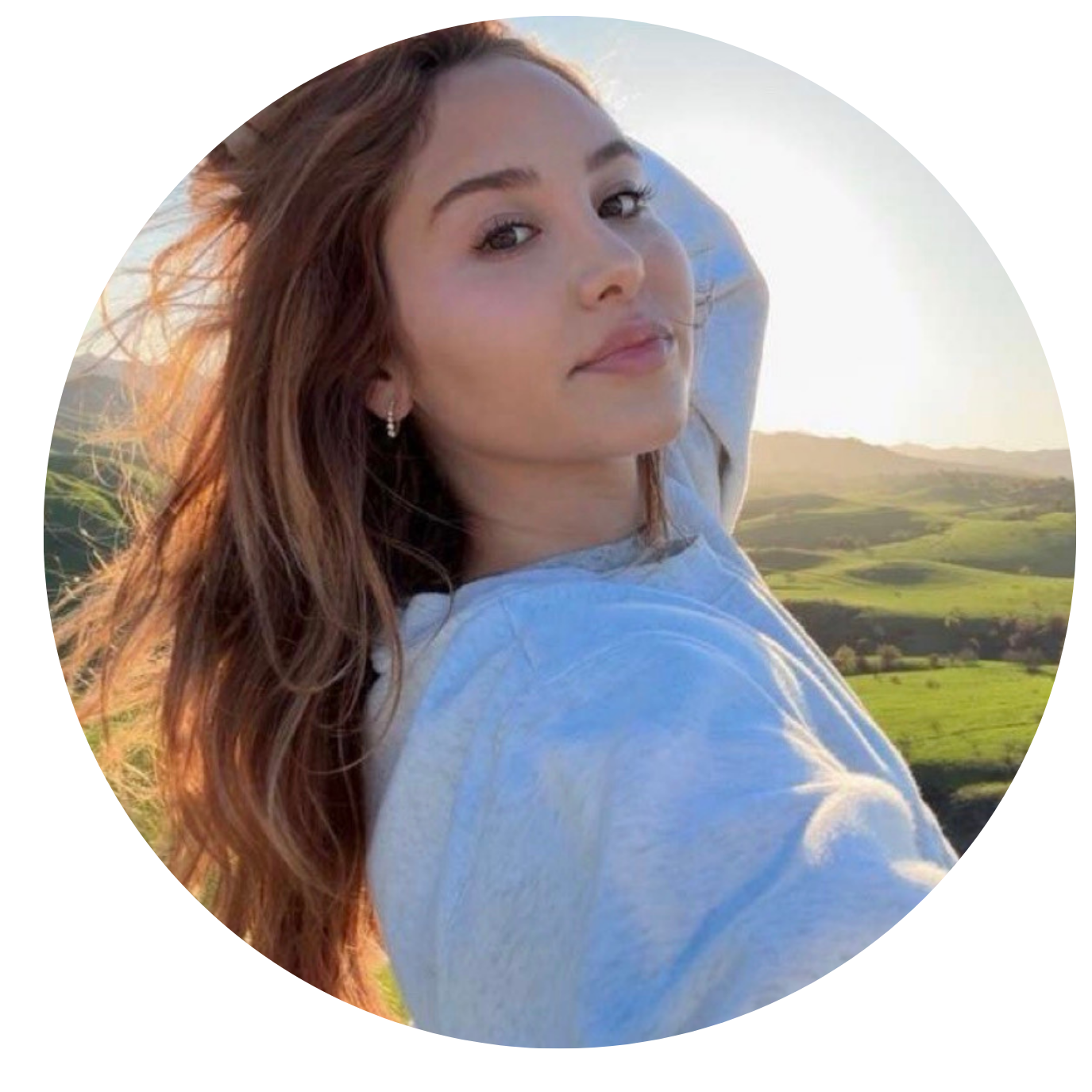 A person outdoors in sunlight, with green fields and hills in the background.