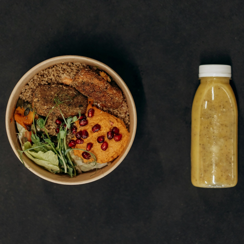 An overhead view of a grain bowl with patties, hummus, and pomegranate seeds next to a yellow smoothie.