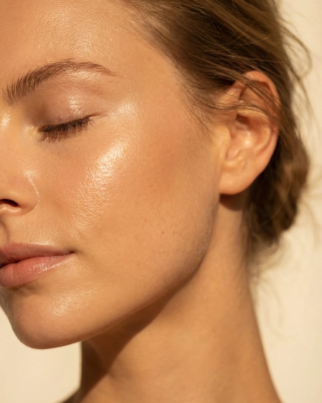 A close-up of a woman's face in profile with her eyes closed, showing glowing, dewy skin.
