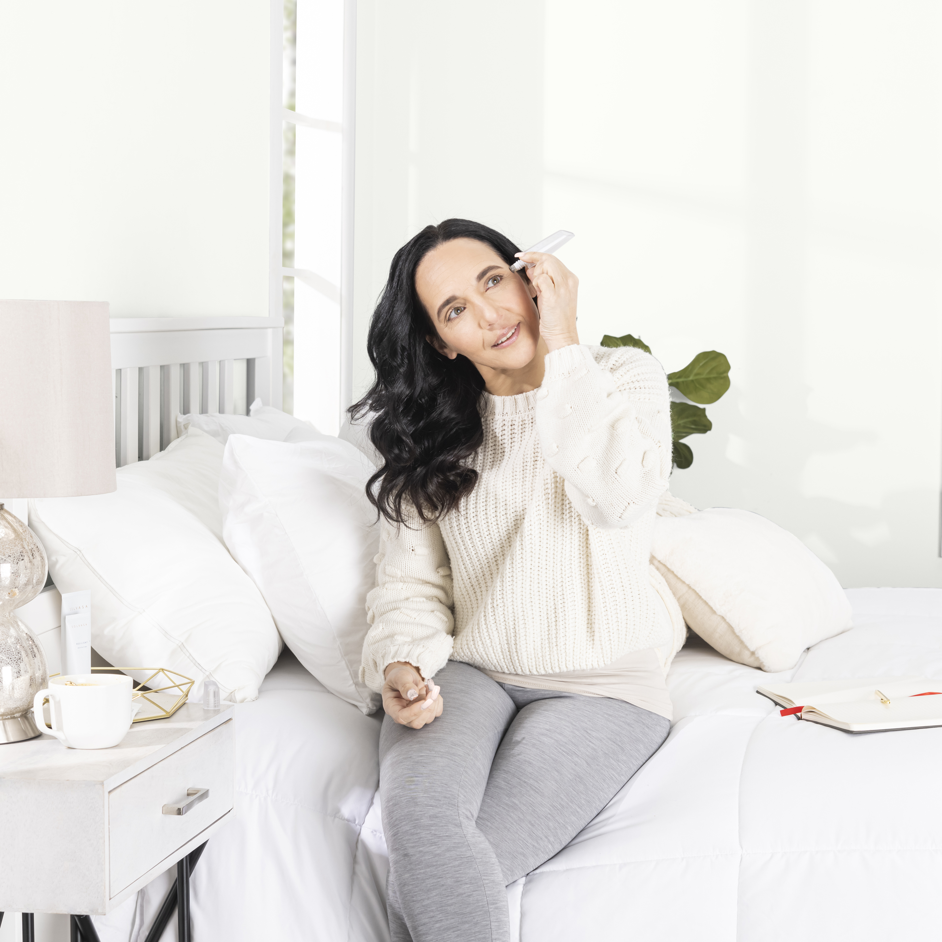 A woman in a sweater sits on a white bed applying a skincare tool to her face.