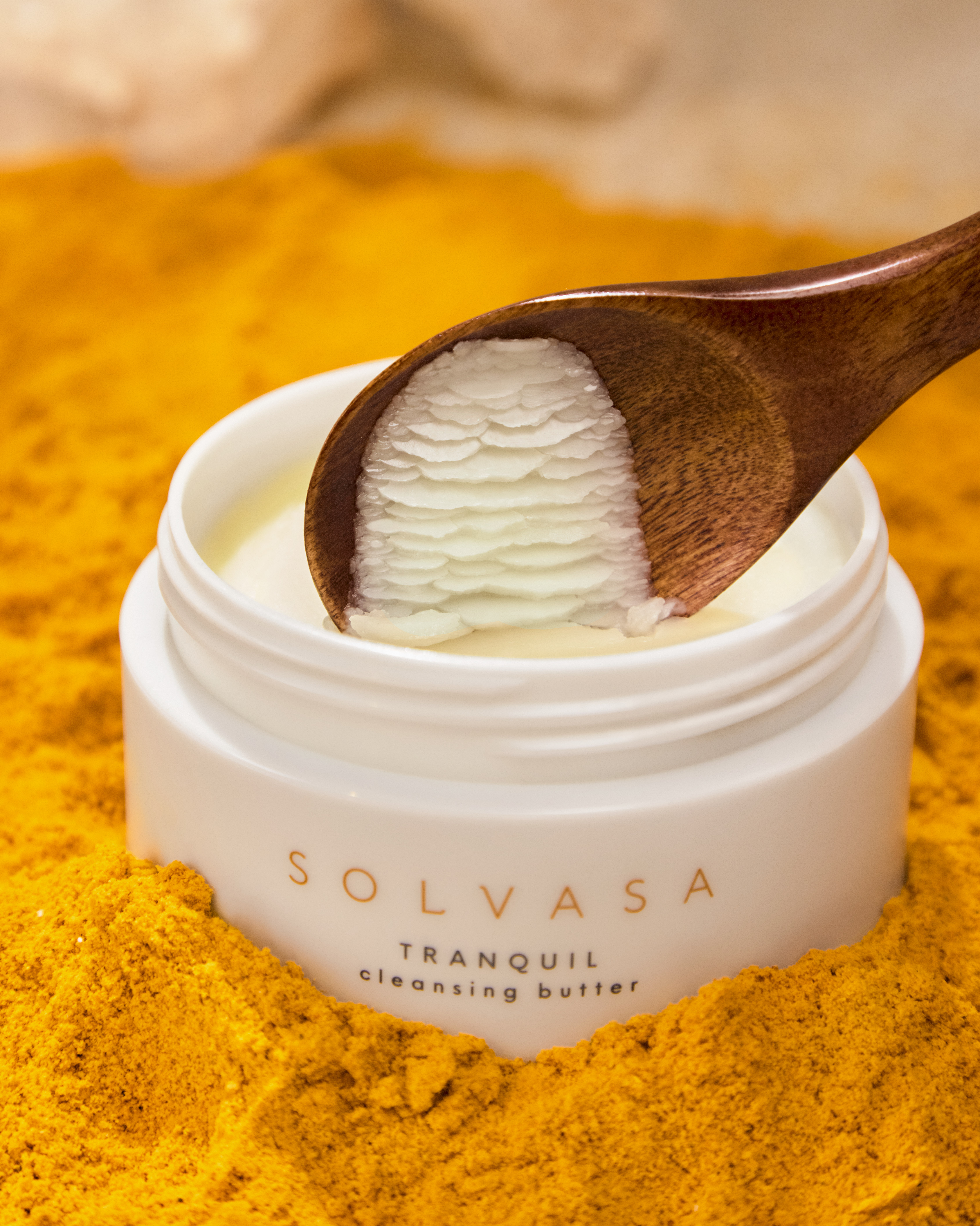 A wooden spoon scoops white cleansing butter from a jar sitting in a bed of yellow powder.