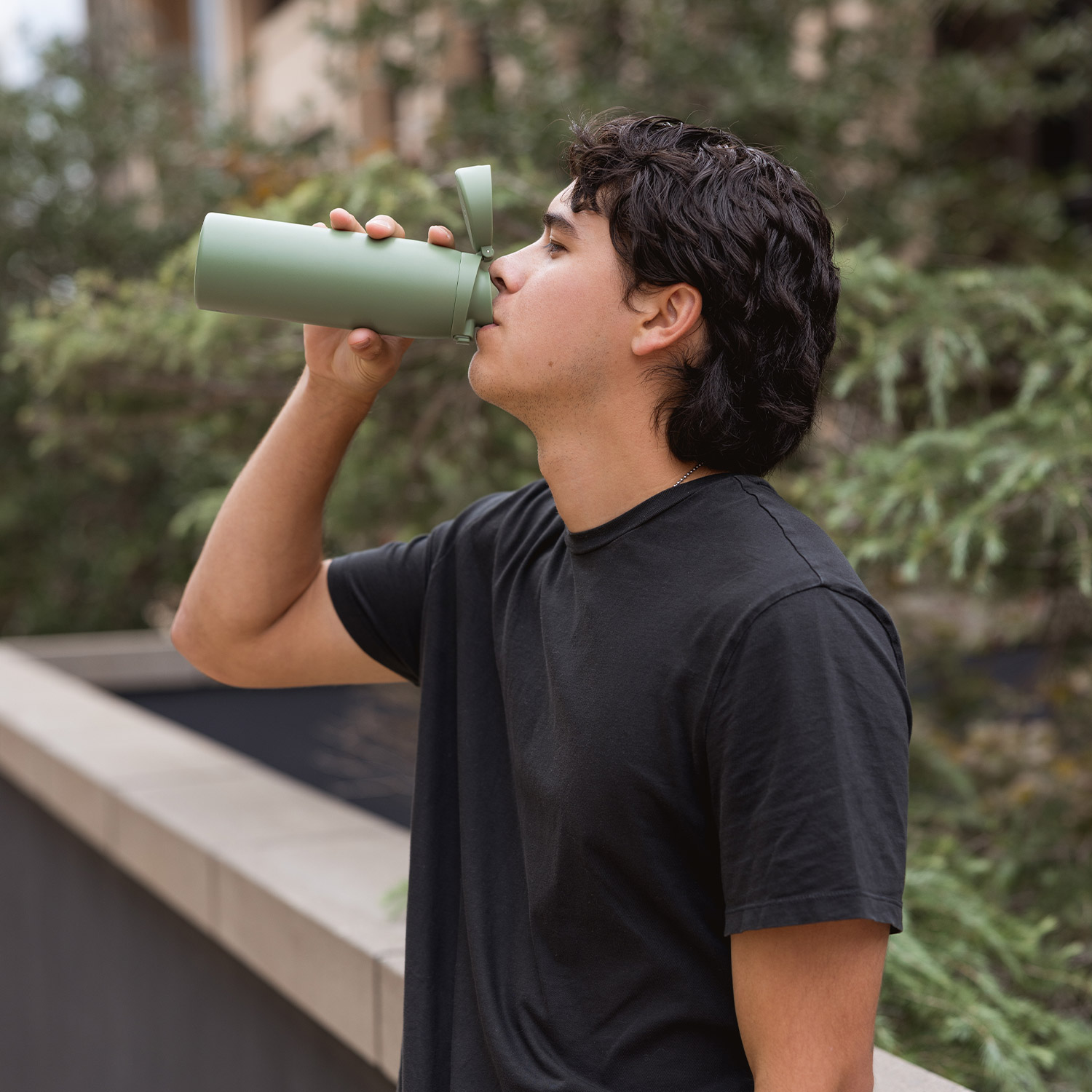 Person in black shirt drinking from a green bottle outdoors.