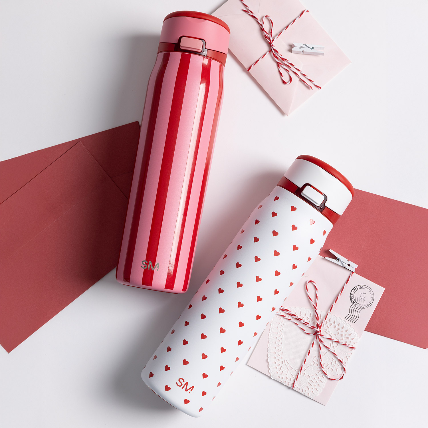 Two Valentine's themed travel mugs, one with stripes and one with hearts, next to red and pink envelopes.