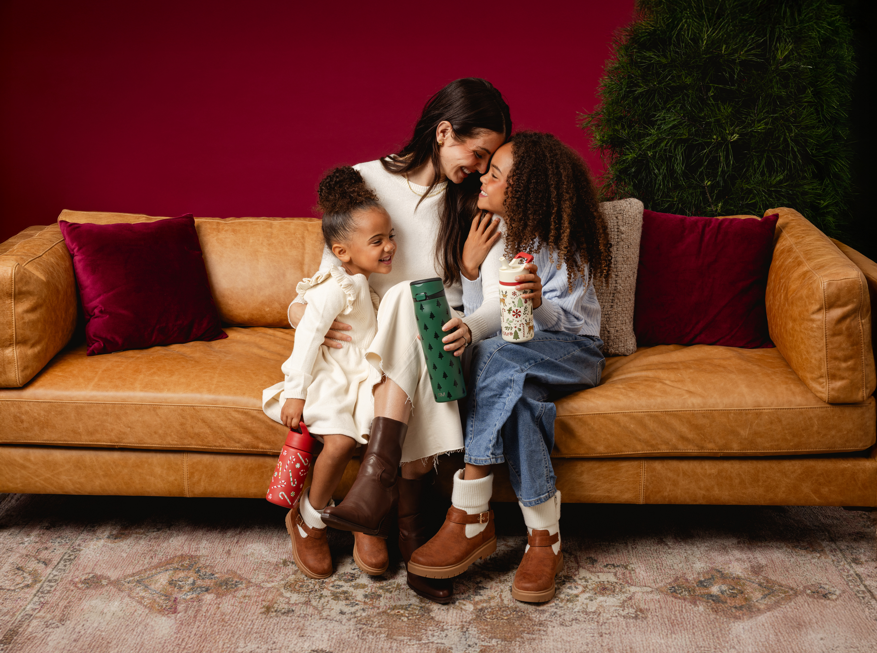 Three people sitting on a couch with colorful pillows, holding festive tumblers.