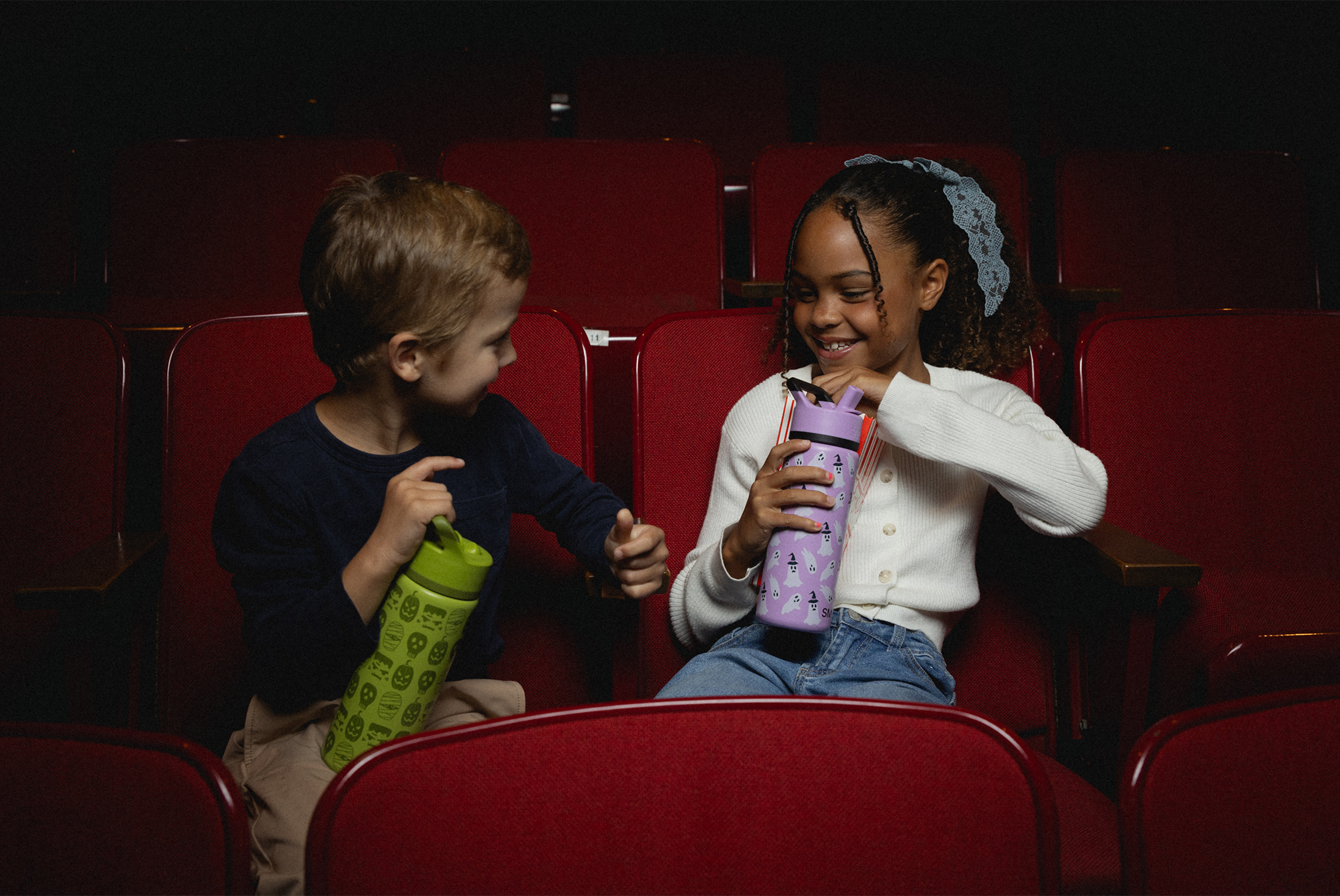 Two children sitting in red theater seats, holding colorful water bottles.