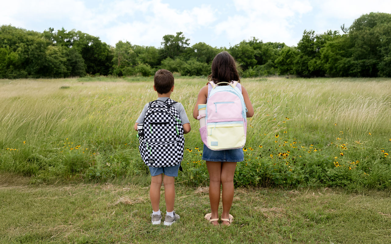 A boy and a girl wearing backpacks stand in a grassy field, seen from behind.